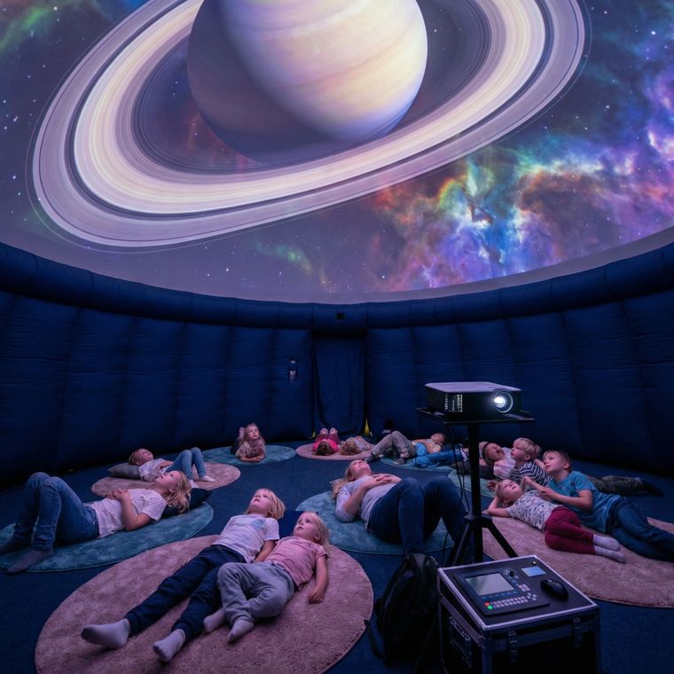 Группа детей лежит на мягких ковриках внутри мобильного планетария, глядя на проекцию планеты Сатурн с кольцами и красочную туманность на куполе